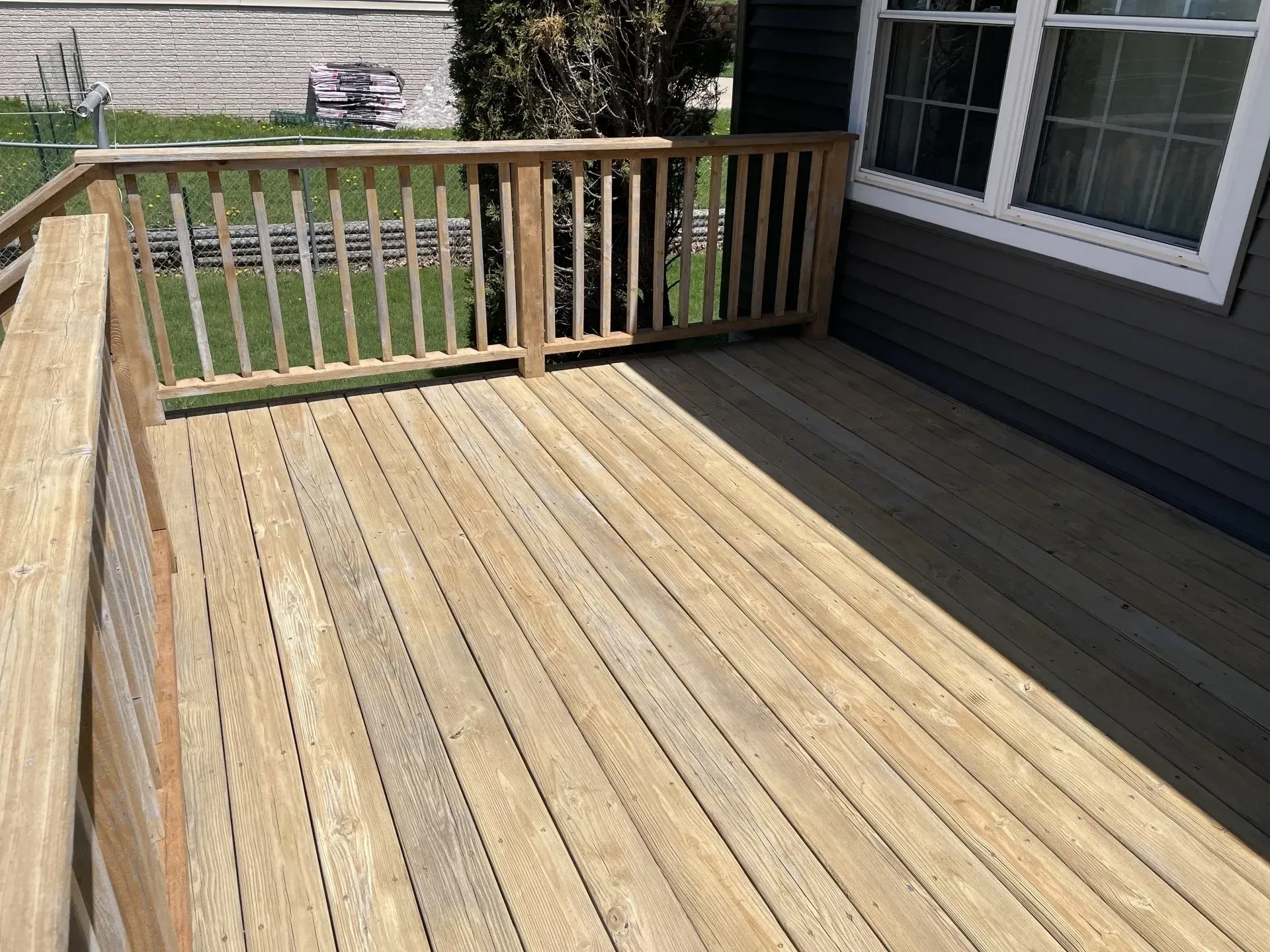A wooden deck with a railing, featuring a clear view of a grassy yard next to a house with dark siding and white windows.