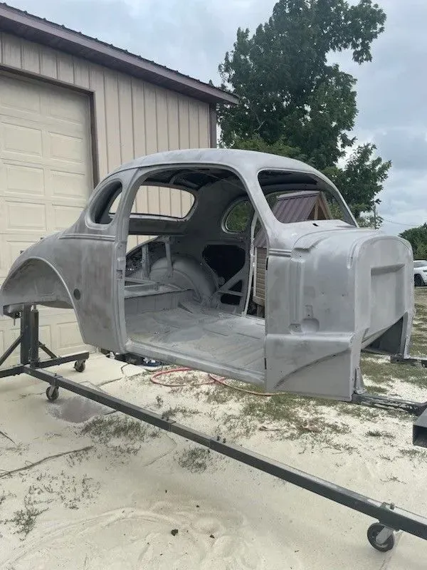 A stripped, primer-gray vintage car body mounted on a metal shop stand outside a building.