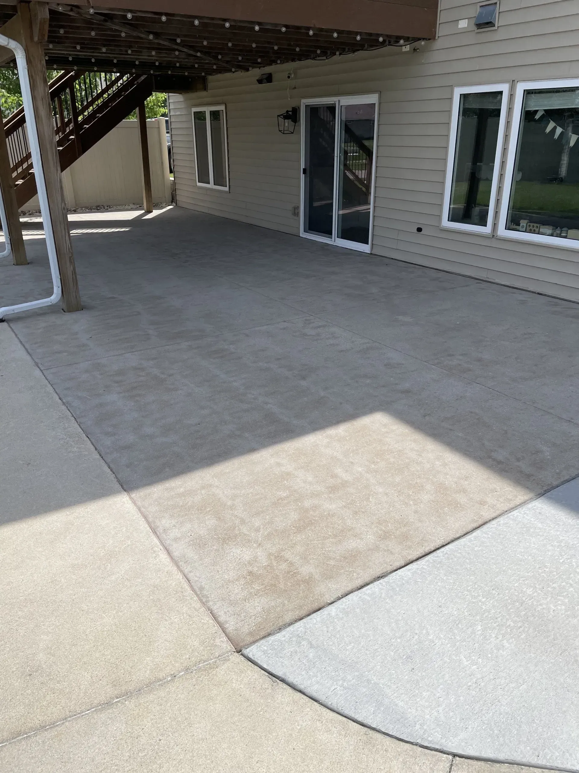 A concrete patio with a wooden staircase and deck overhead, adjacent to the side of a house with sliding glass doors.