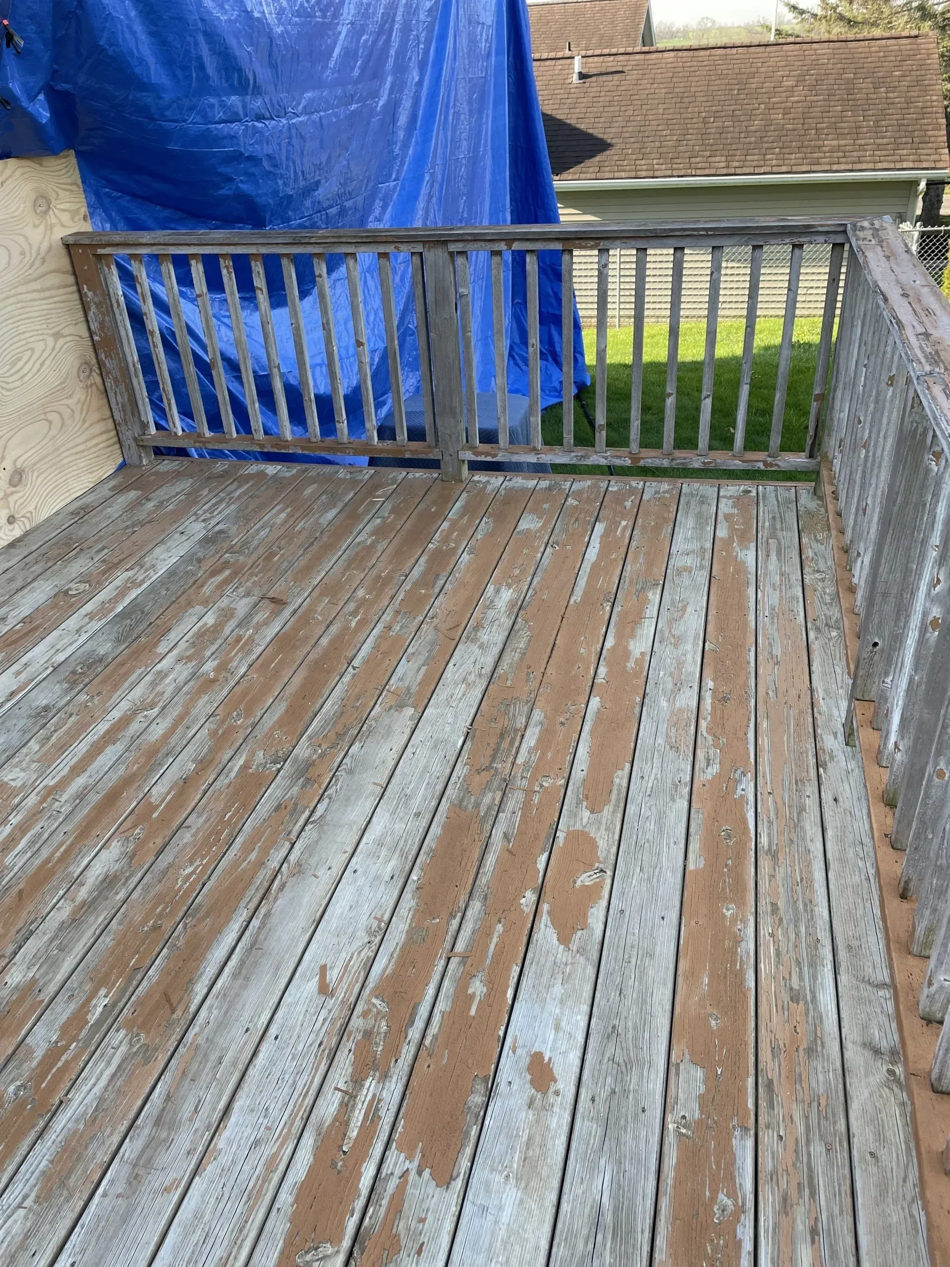 An outdoor wooden deck with weathered, peeling gray paint, featuring a simple railing and a blue tarp in the background.