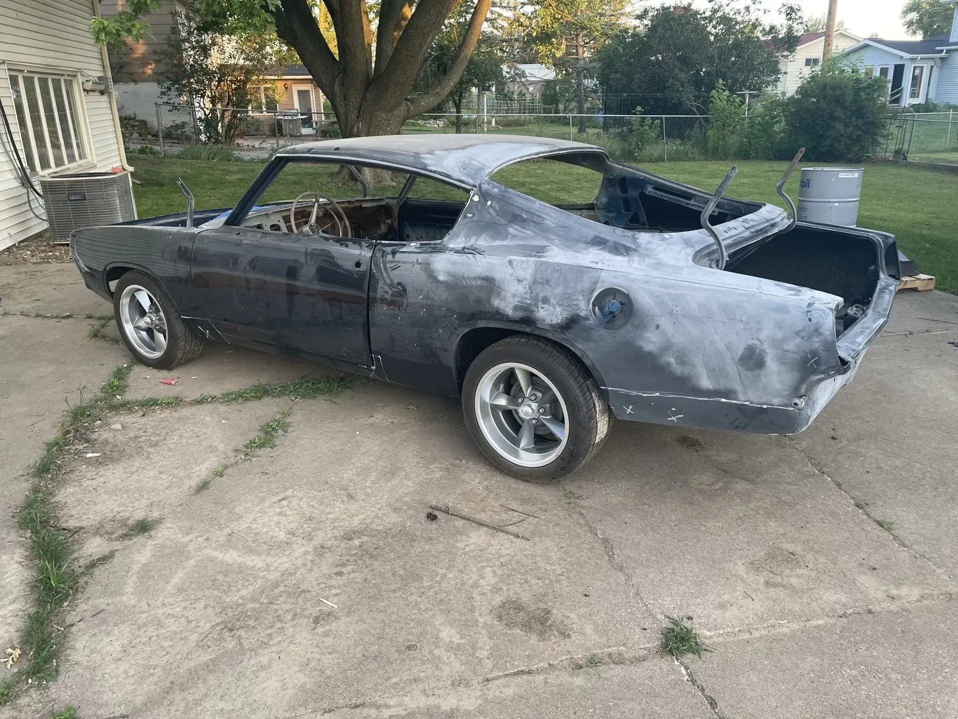 A partially sanded black muscle car project parked on a concrete driveway with a house and trees in the background.