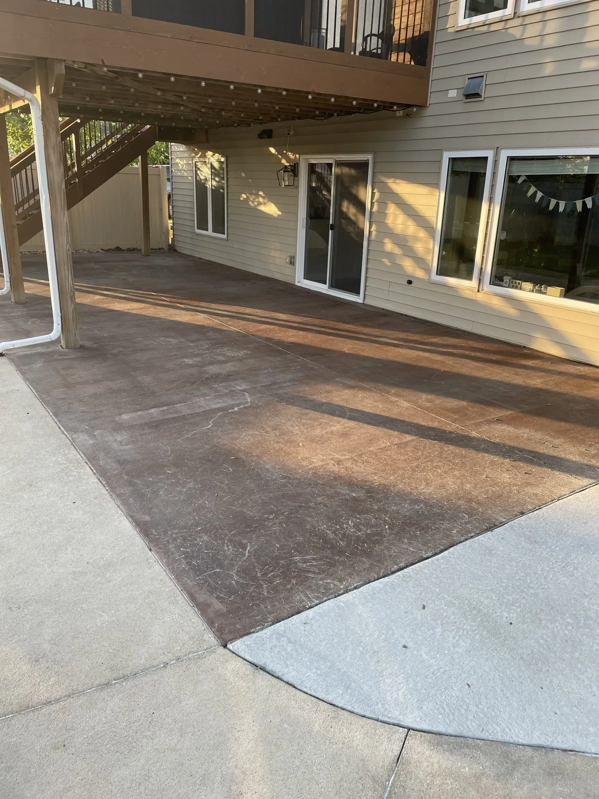 A large, multi-textured concrete patio under a wooden deck with a sliding glass door and windows in the background.