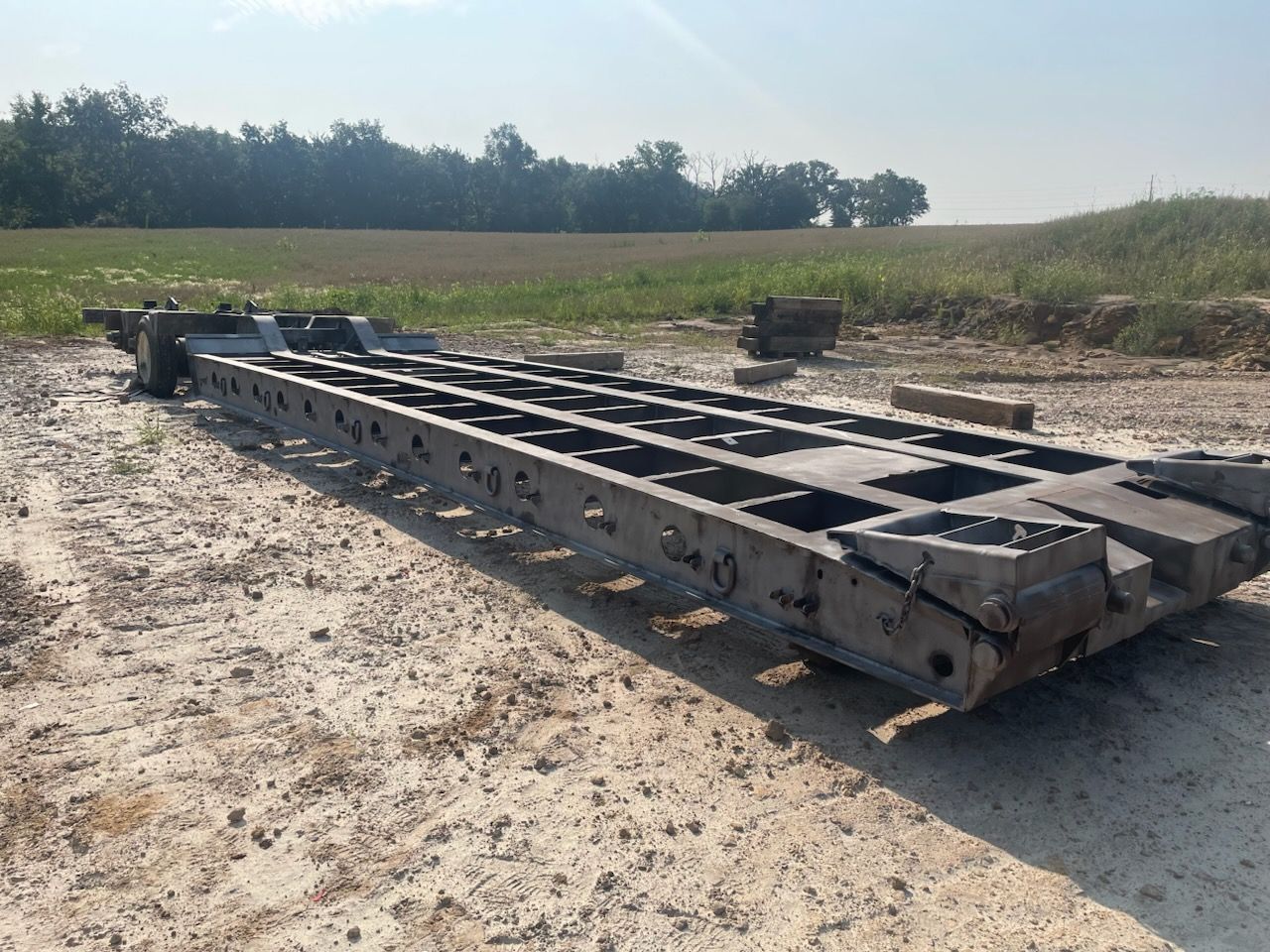 A long, empty metal trailer frame sits on a dirt lot in front of a field and tree line under a clear, bright sky.