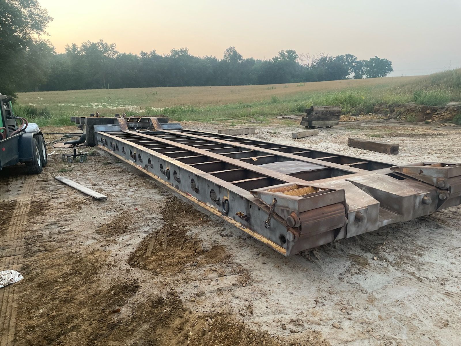 A heavy-duty steel lowboy trailer frame rests on a dirt lot with a vehicle visible on the left.
