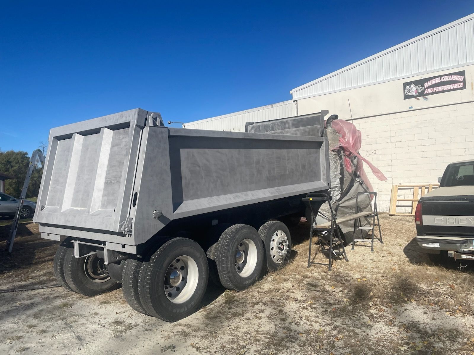 A grey, heavy-duty lowboy trailer with a detachable gooseneck parked on a dirt lot under a cloudy sky.