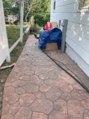 A stamped concrete walkway leads alongside a white house with a blue-tarped object and two orange cones resting on it.