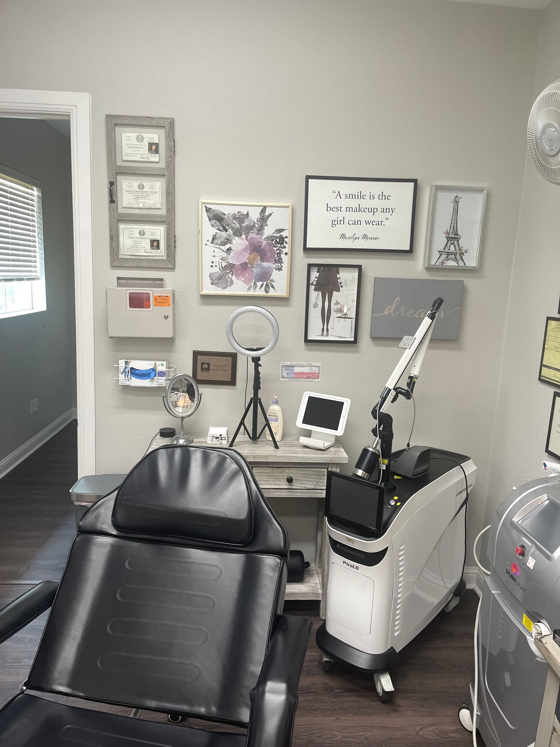 A medical room with a black reclining chair, laser machine, and framed art on a gray wall.