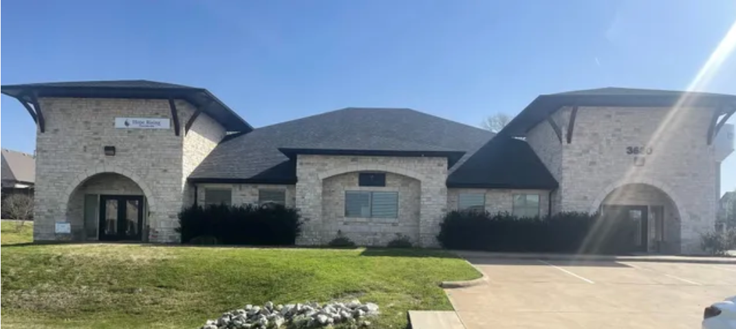 Exterior of a light stone-colored office building under a blue sky. Features dark roof and arched entrances.