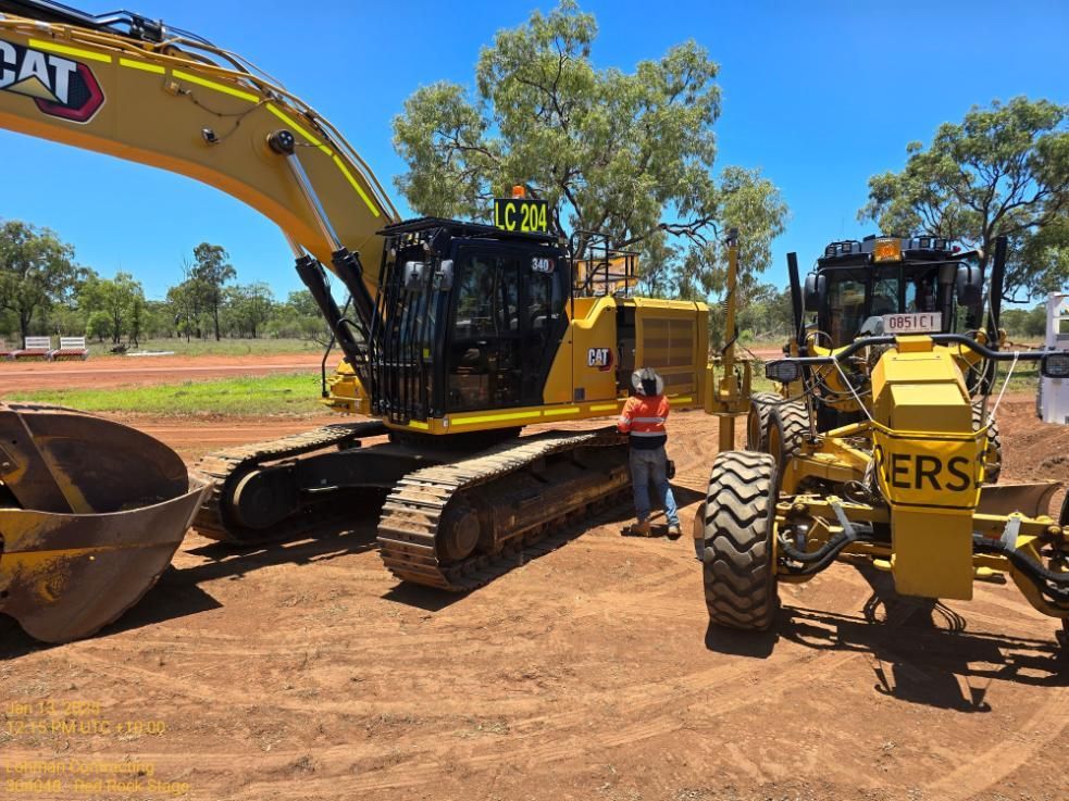 A Man is Standing Next to a Cat Excavator and a Grader — Lohman Contracting in Emerald, QLD