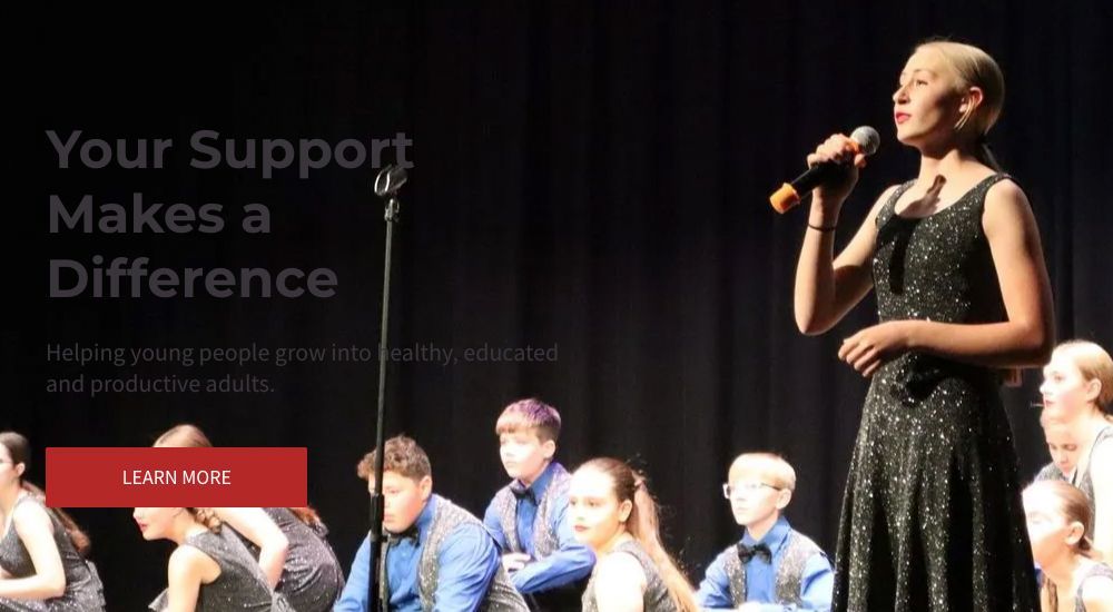 Young woman singing on stage, dressed in black, with a group of young people in the background.
