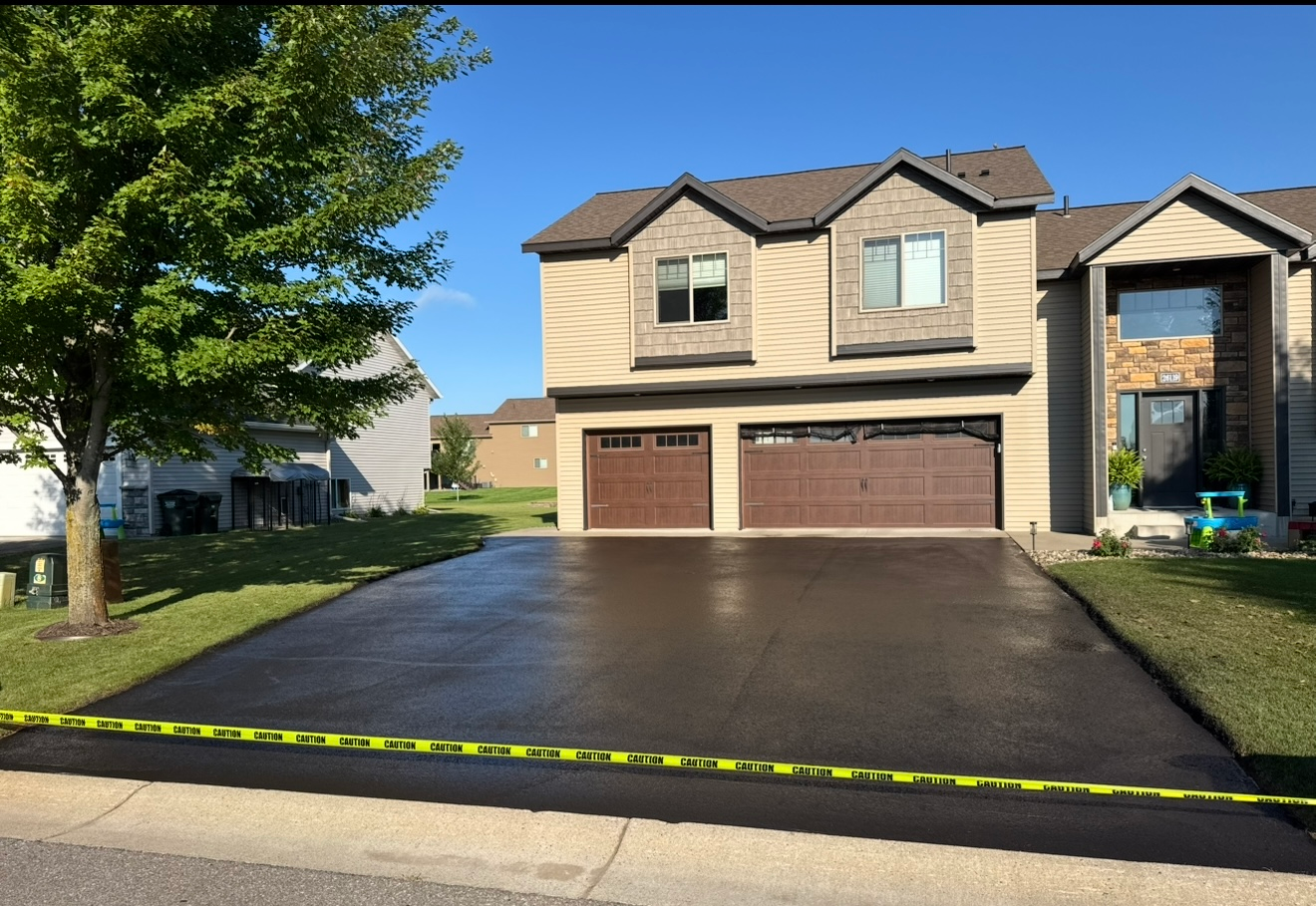Suburban two-story house with a three-car garage and freshly paved driveway in bright sunlight.
