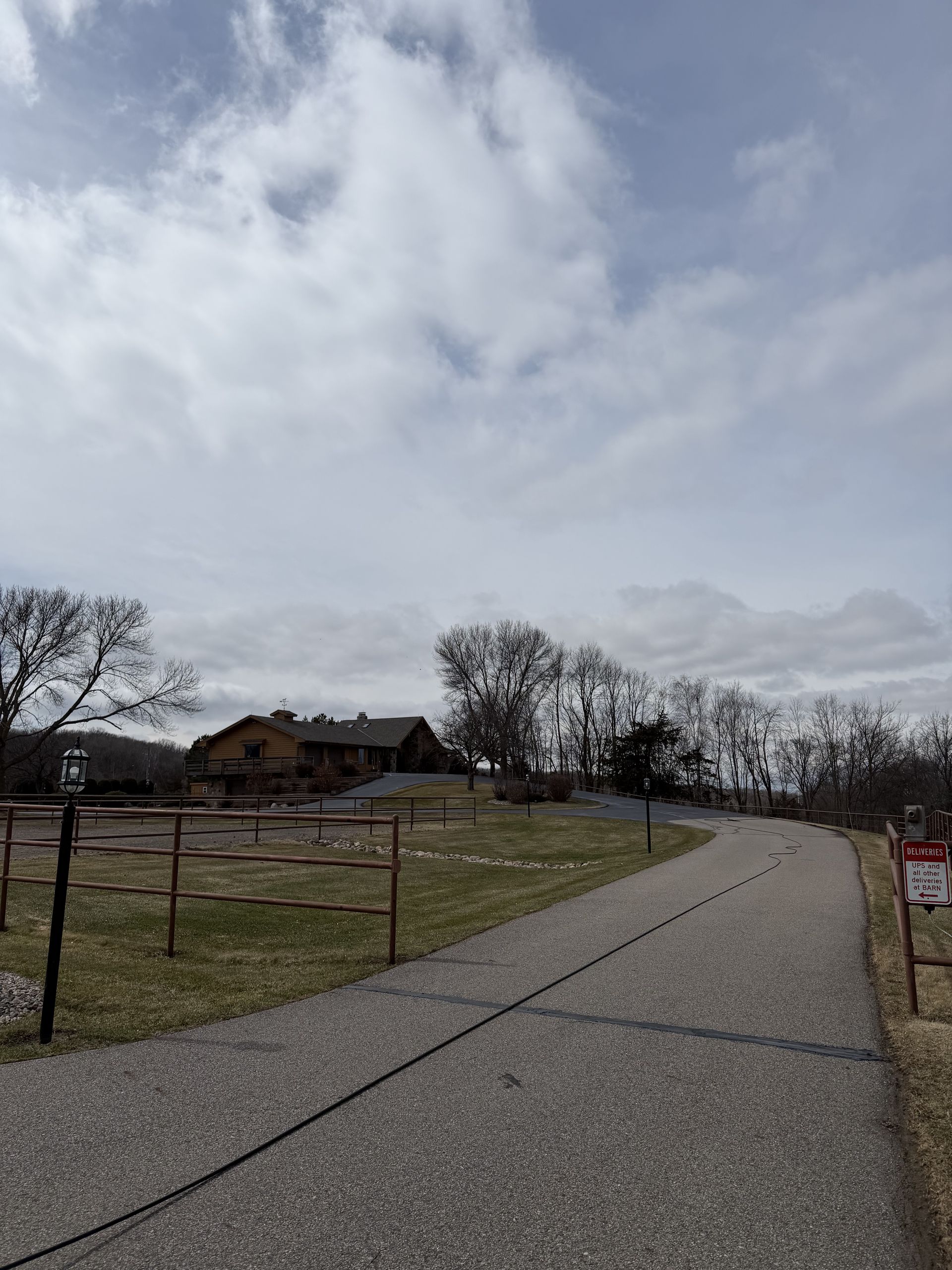 Rural gravel road curving past a fenced field and house under a cloudy sky