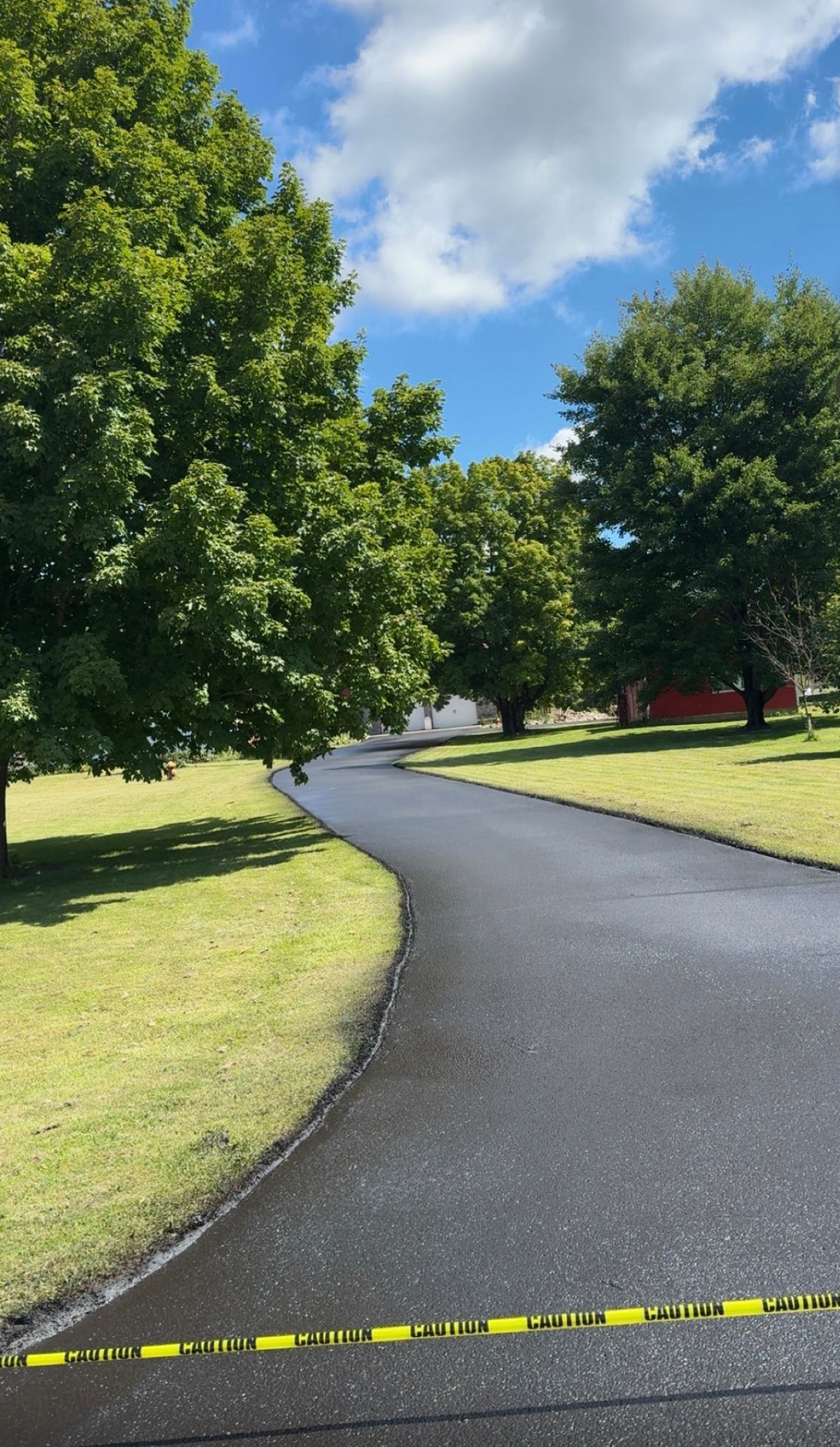 Curving paved path through green trees and grass under a blue sky, with yellow caution tape in the foreground