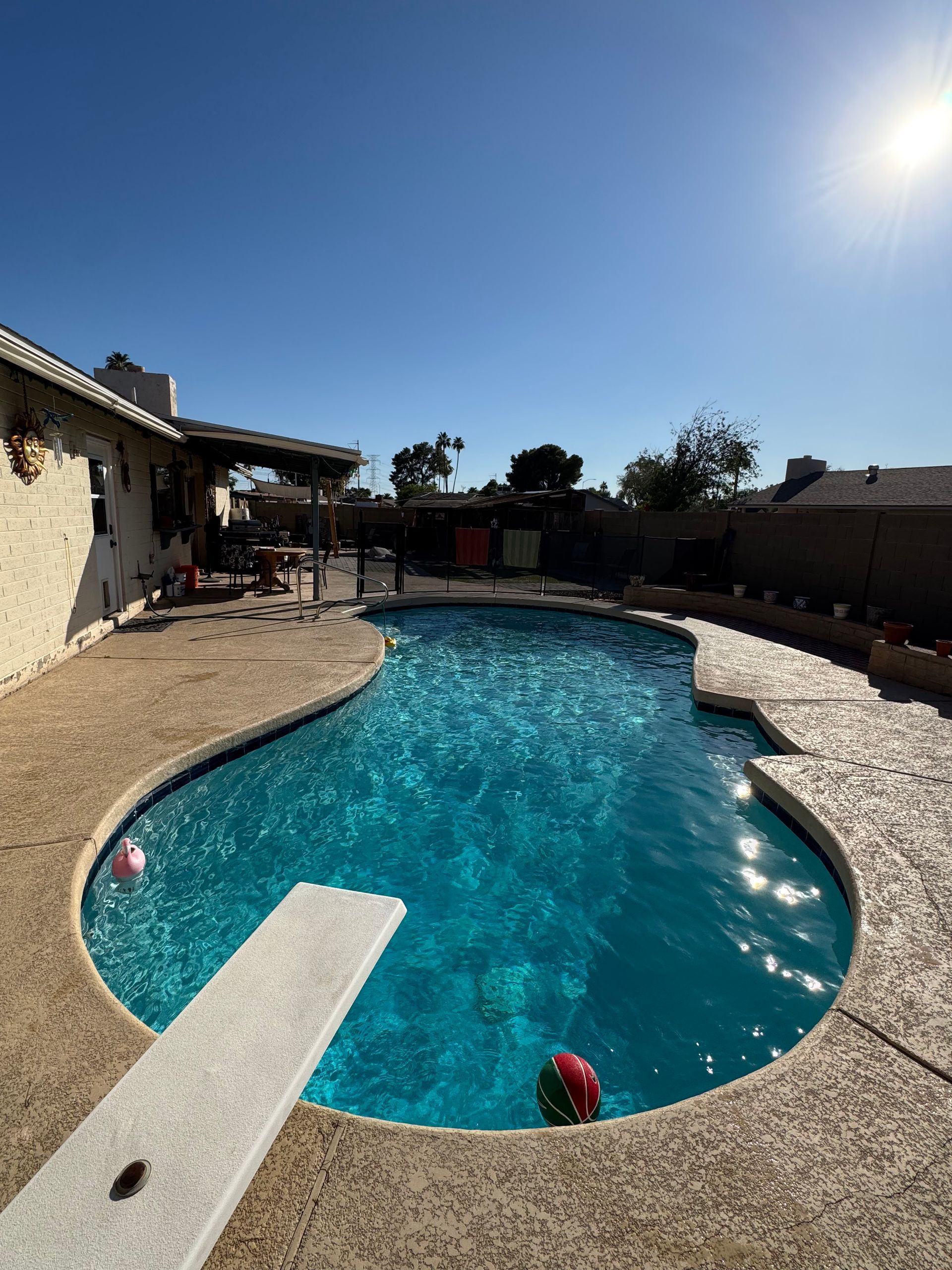 A backyard pool on a sunny day, with a diving board and two inflatable balls.
