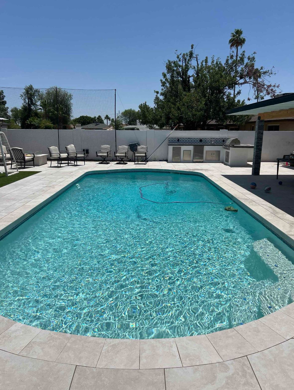 Swimming pool with blue water surrounded by light-colored stone. Outdoor setting with chairs, grill, and blue sky.