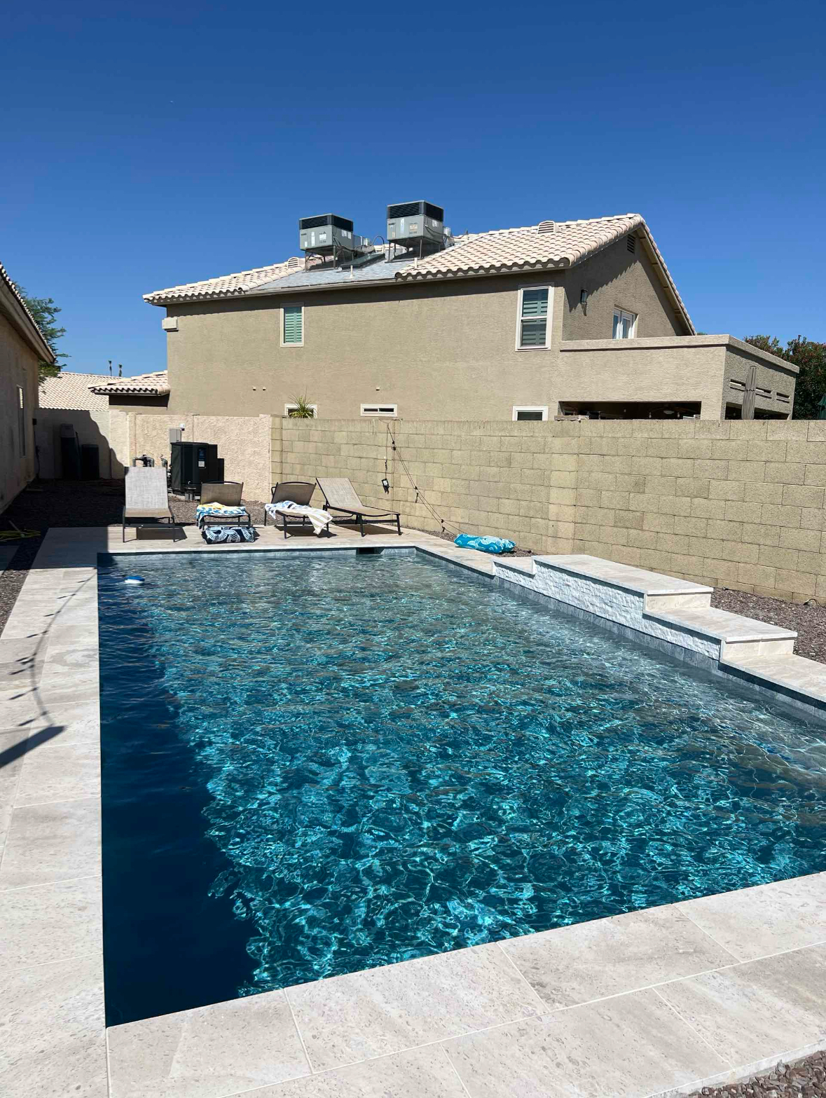 Rectangular swimming pool in a backyard, with a house and a wall. Sunny day, blue water.