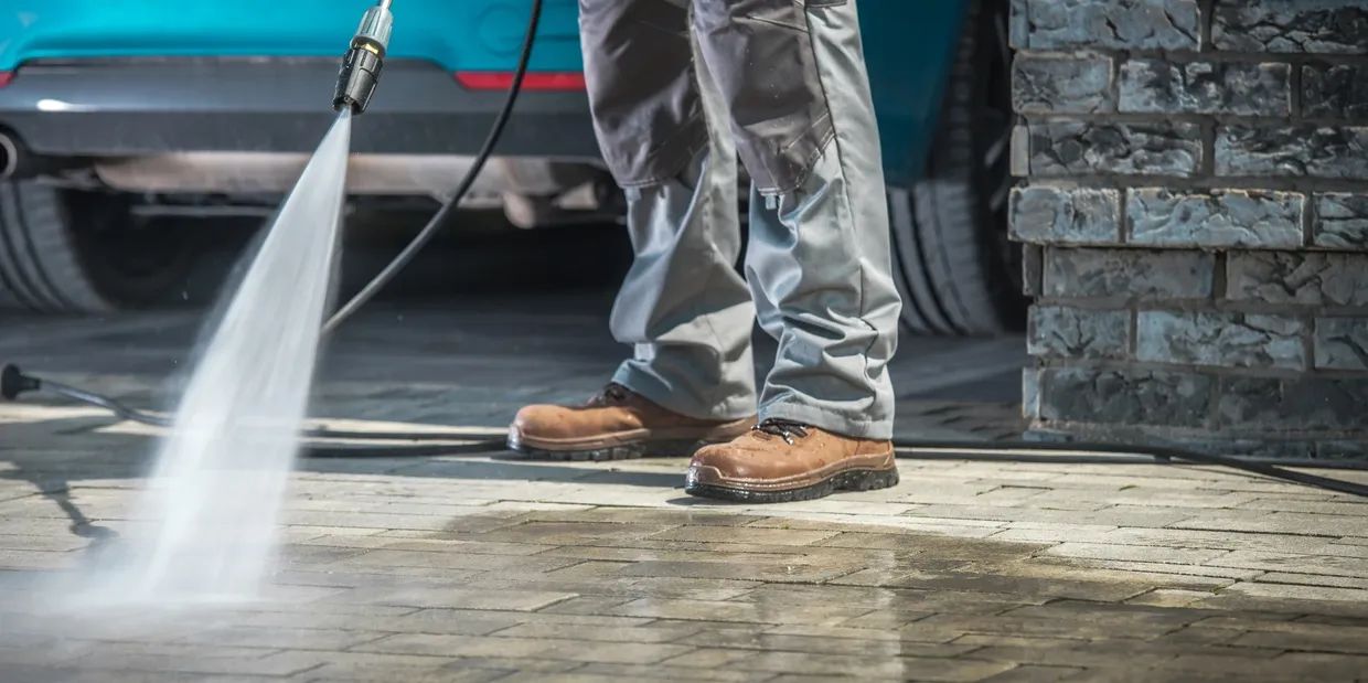 Person power washing a concrete surface near a teal car and brick wall.
