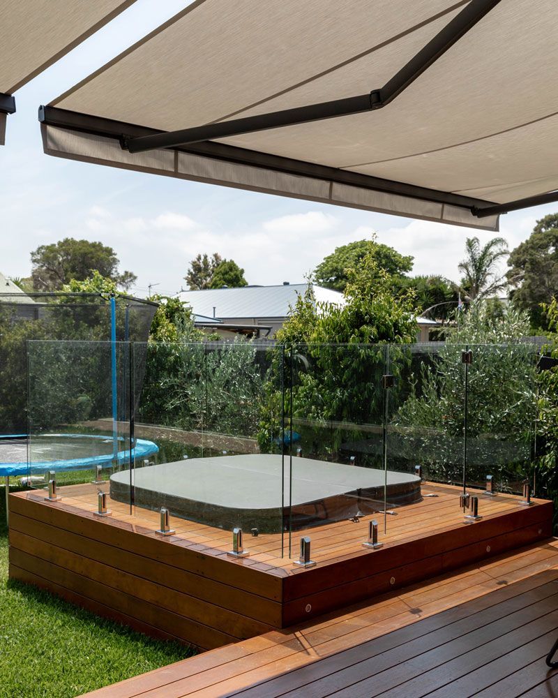 Wooden deck with hot tub, glass railing, and retractable awning in a backyard.