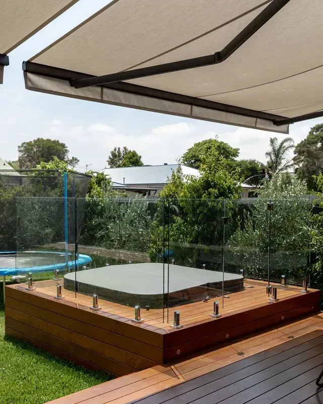 Wooden deck with hot tub, glass railing, and retractable awning in a backyard.