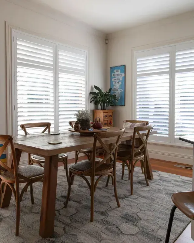 Dining room with wooden table, chairs, and shutters. A blue artwork and plants decorate the space.