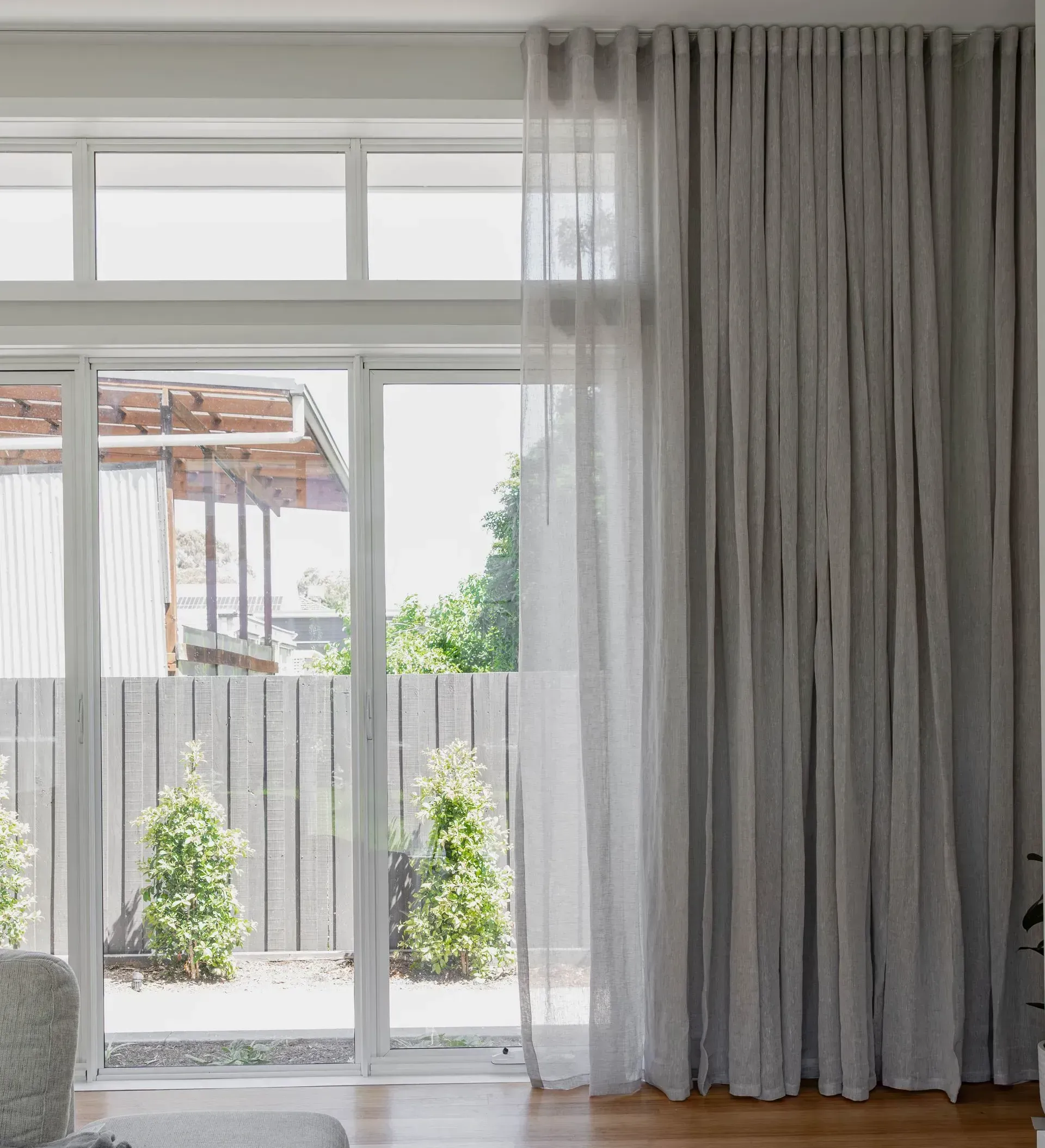 Sheer gray curtains framing a bright window, looking out to a backyard with trees and a shed.