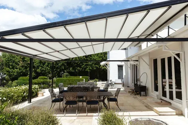 Patio with a dining table under a white canopy with black beams. White house in the background, sunny outdoor setting.