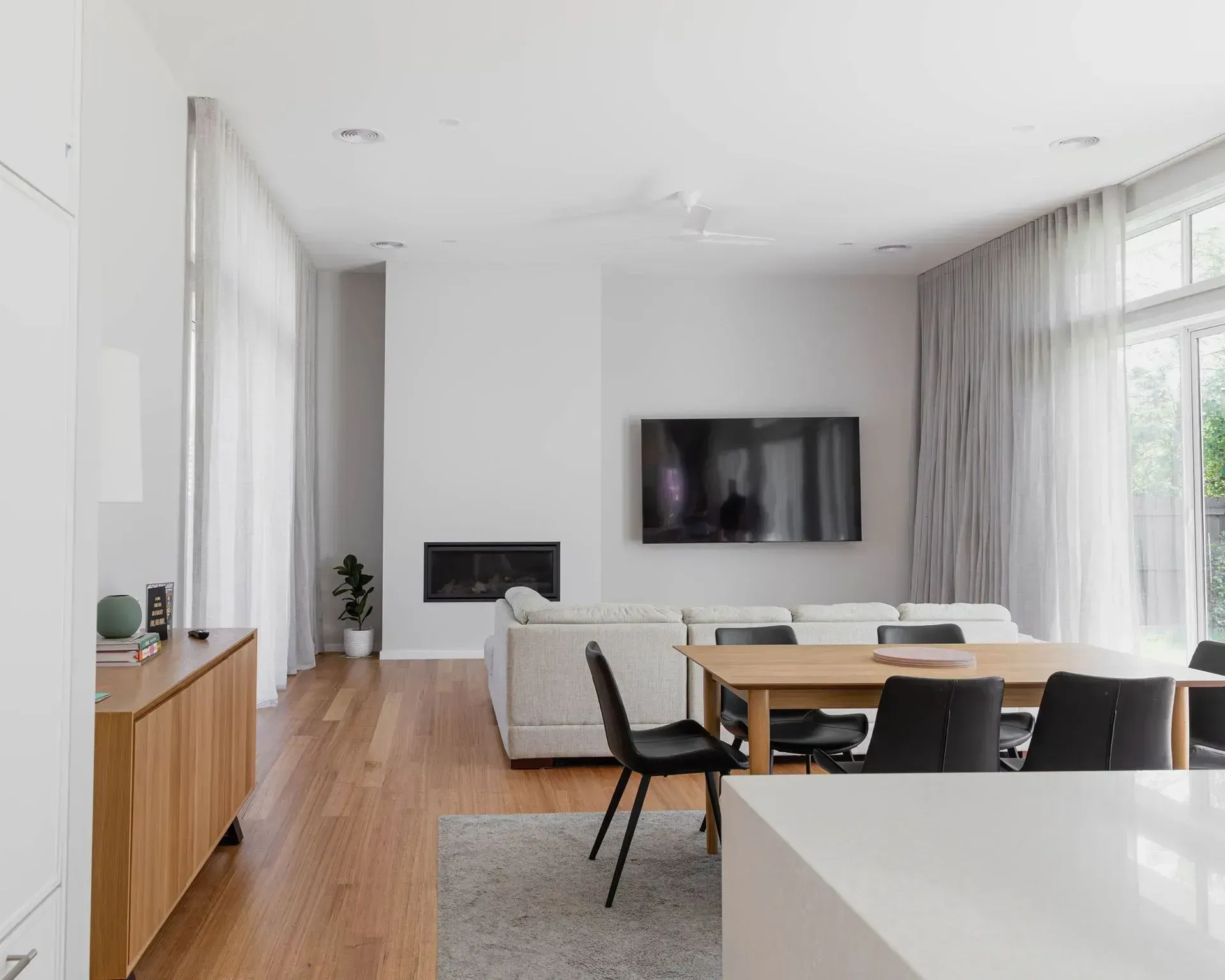 Spacious living room with wood floors, white walls, and a dining table, featuring a fireplace, TV, and sheer curtains.