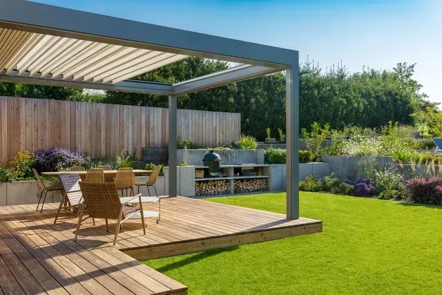 Wooden patio with dining set, grill, and pergola in a sunny garden.