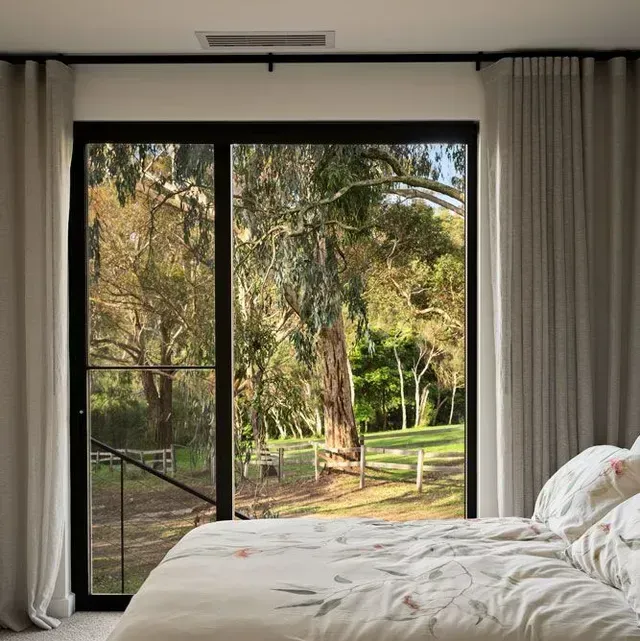 Bedroom with large window overlooking a lush green yard; beige curtains frame the view, a bed is in the foreground.