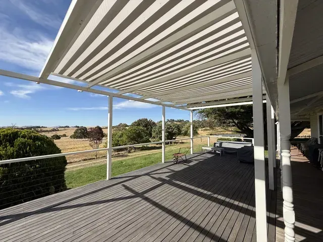 White-framed patio with striped roof, casting shadows on a wooden deck overlooking a rural landscape under a blue sky.