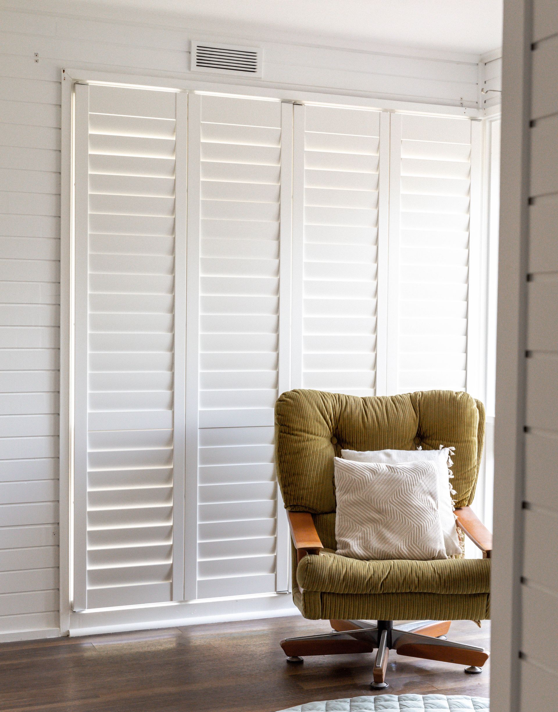 Bedroom with white shutters, round chandelier, and a blue bedspread. A chair sits to the left.