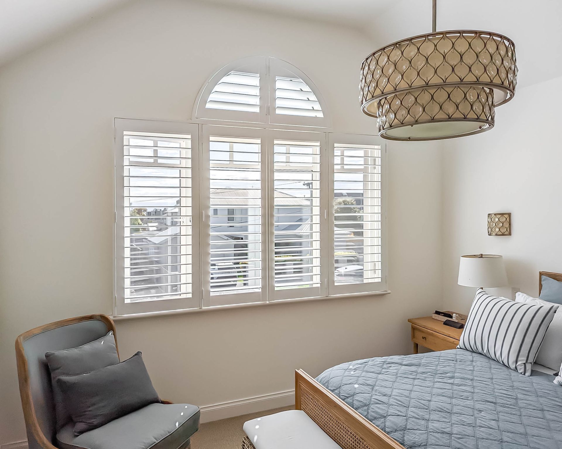 Bedroom with white shutters, round chandelier, and a blue bedspread. A chair sits to the left.