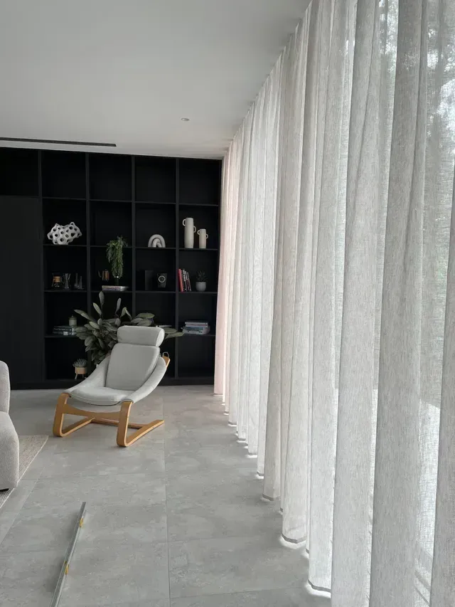 Modern living room with sheer white curtains, a chair, and a black bookshelf with decor.