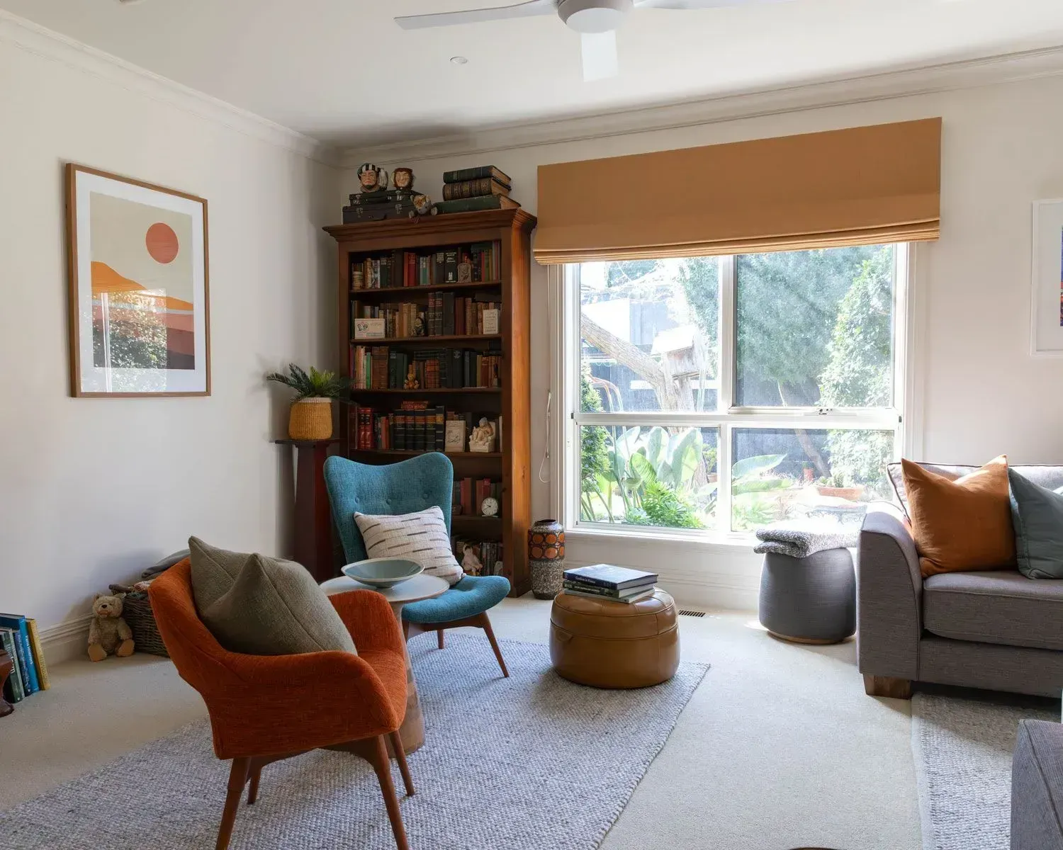 Living room with orange and teal chairs, bookshelf, window with tan shade, and a gray sofa.