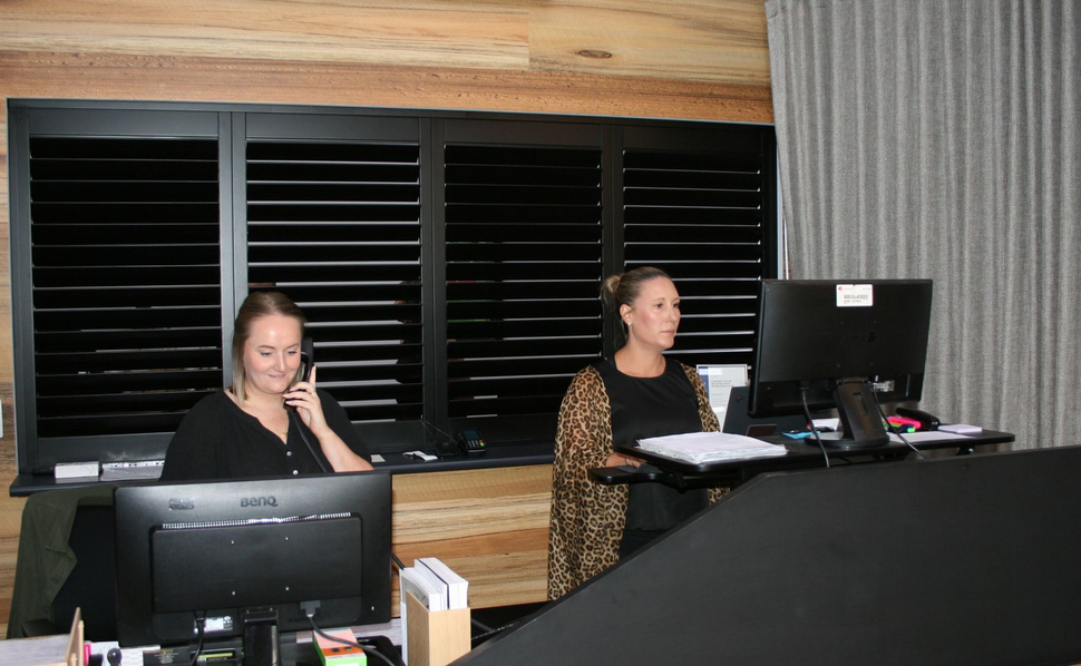 Two women working at desks with computers, behind a black counter, inside a wood-paneled office.