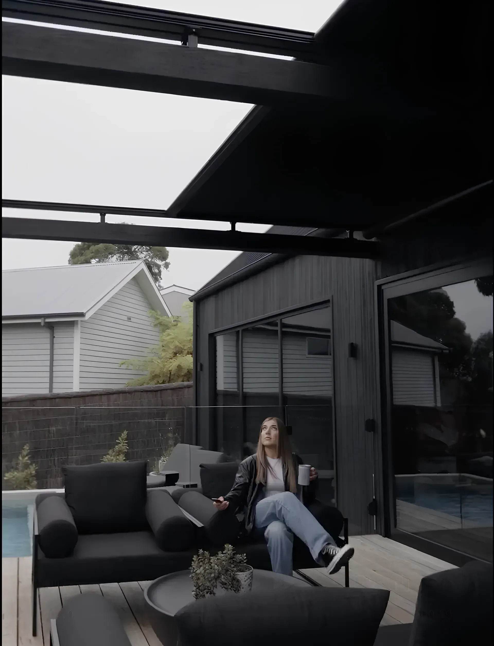 Woman relaxing on outdoor sofa, holding a mug. Black patio furniture and overhead awning.