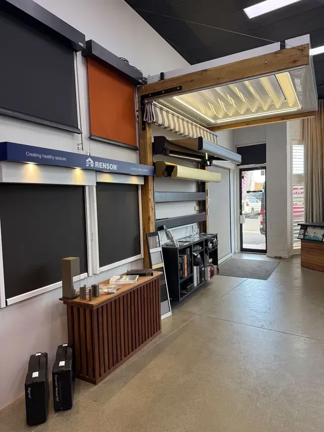 Interior of a showroom displaying retractable awnings and blinds. Various colors are shown, with a wooden cabinet and shelves.
