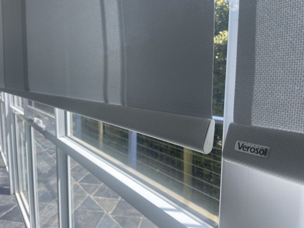 Roller shades partially cover a window. Light gray fabric, white frame, outdoors visible. 