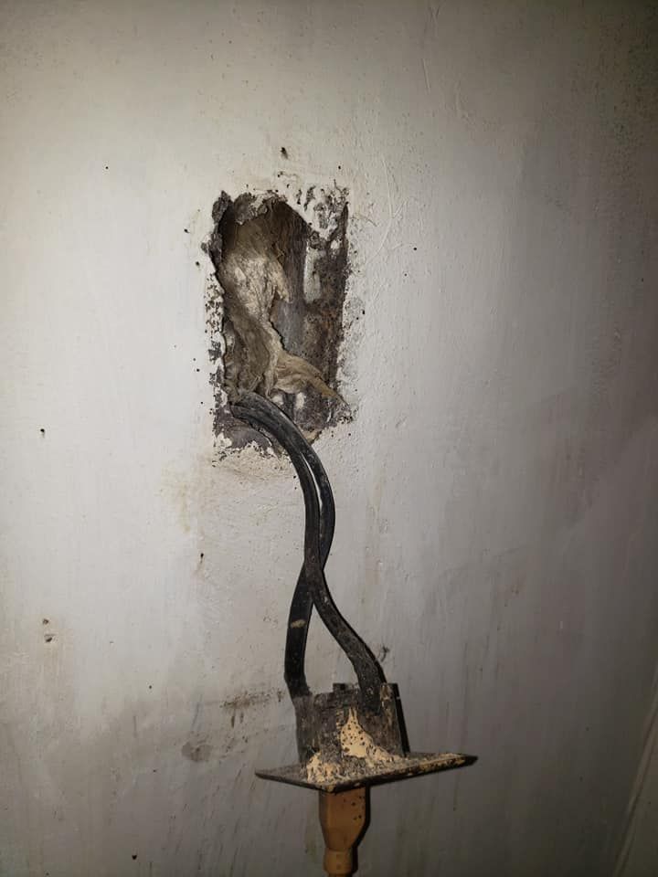 Damaged electrical outlet in a wall, with exposed wires and crumbling plaster.