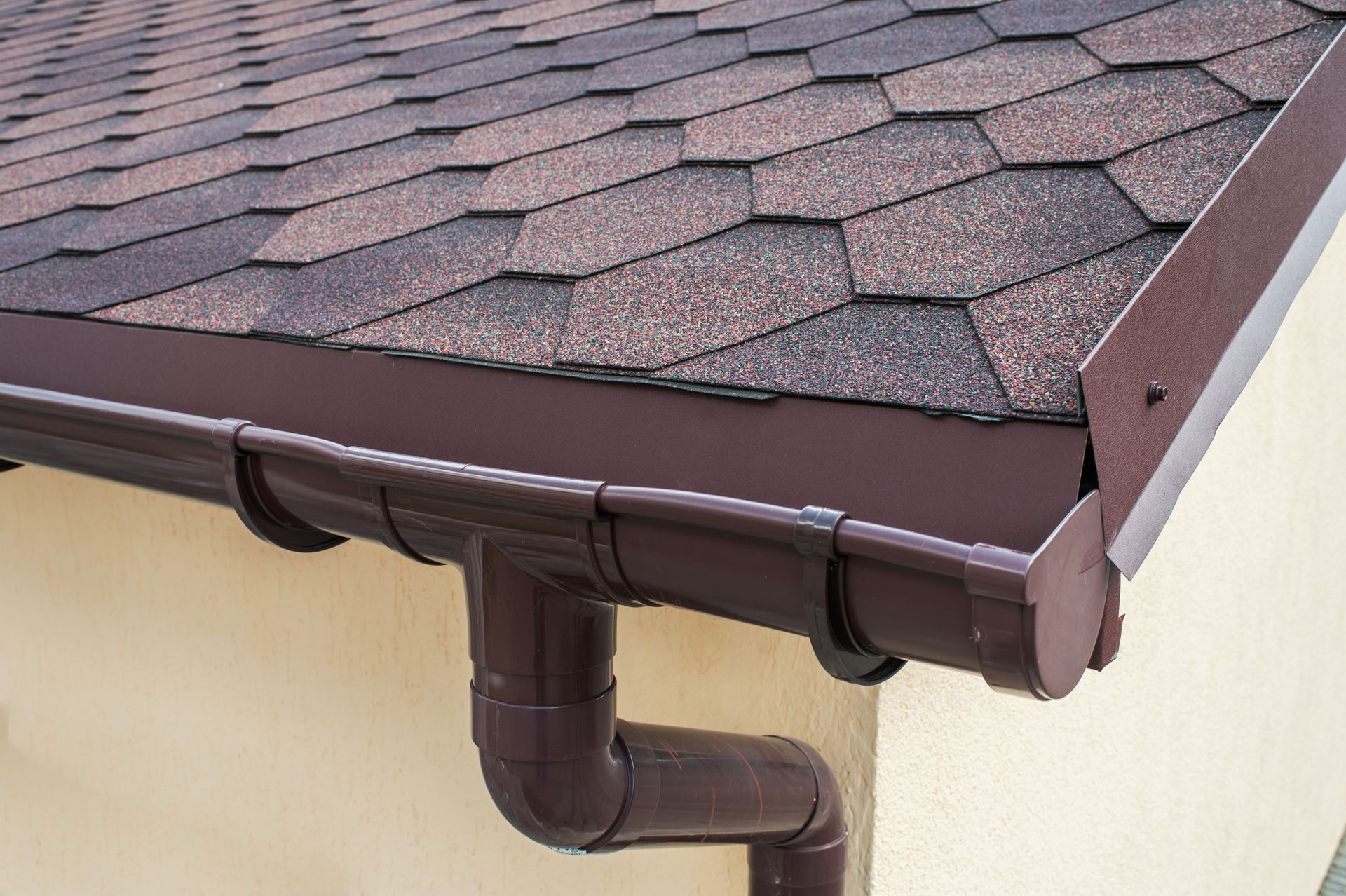 Brown rain gutter and downspout under a shingled roof corner on a beige house