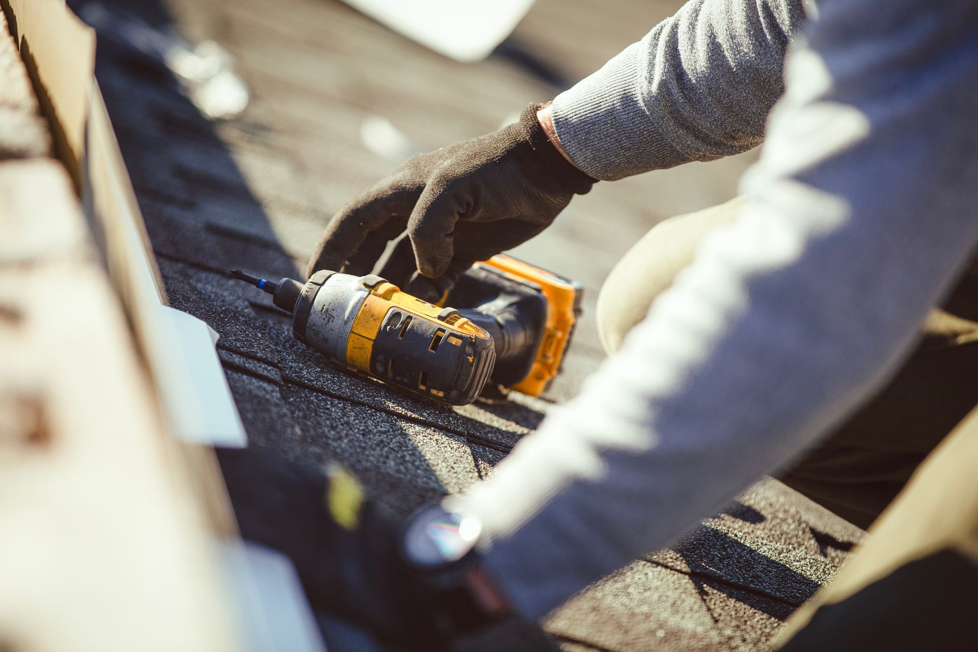 Person in gloves using a power drill on a rooftop, fastening shingles.