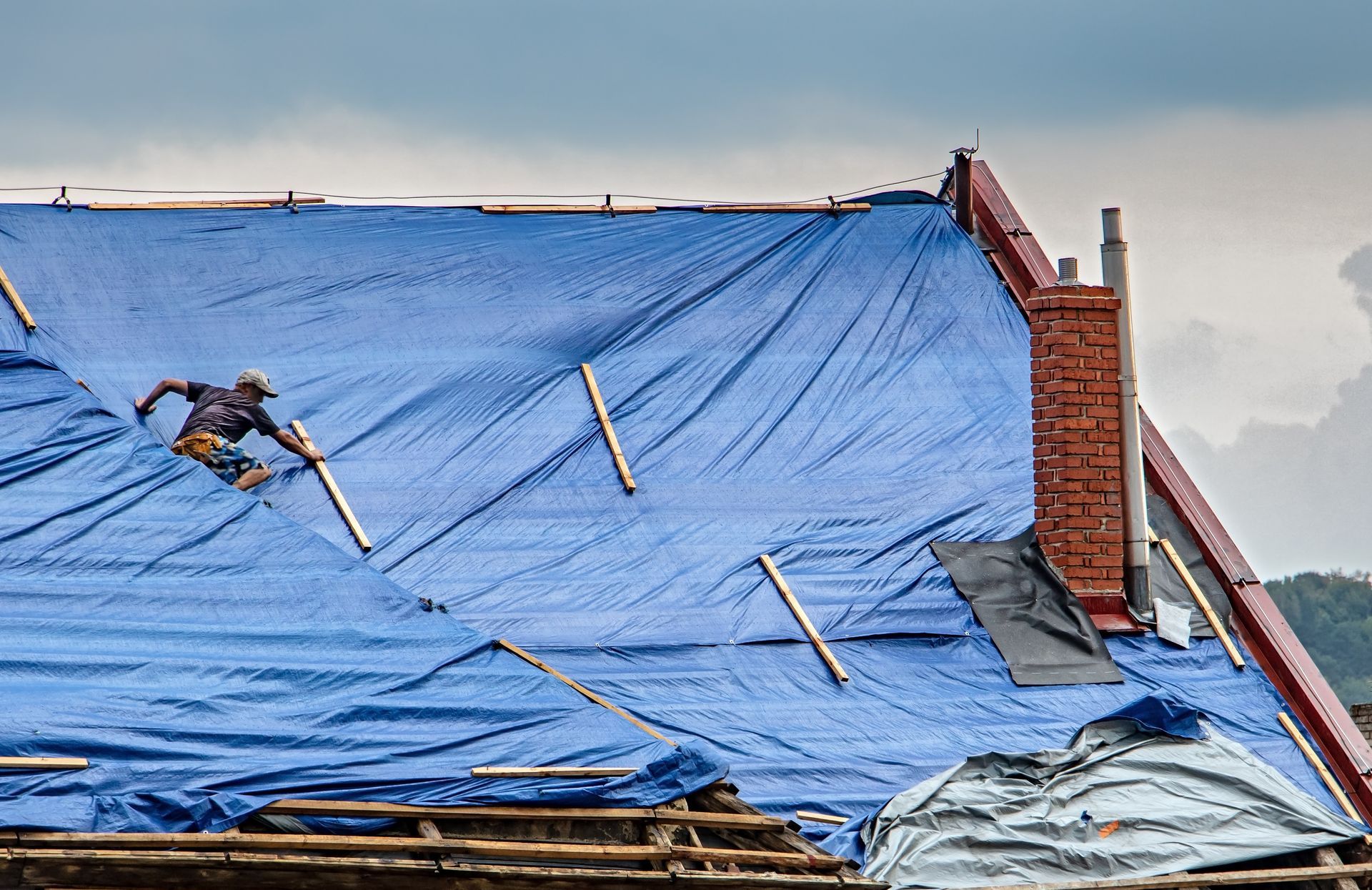 Blue tarp covers damaged roof with two workers on top, near chimney and brick wall