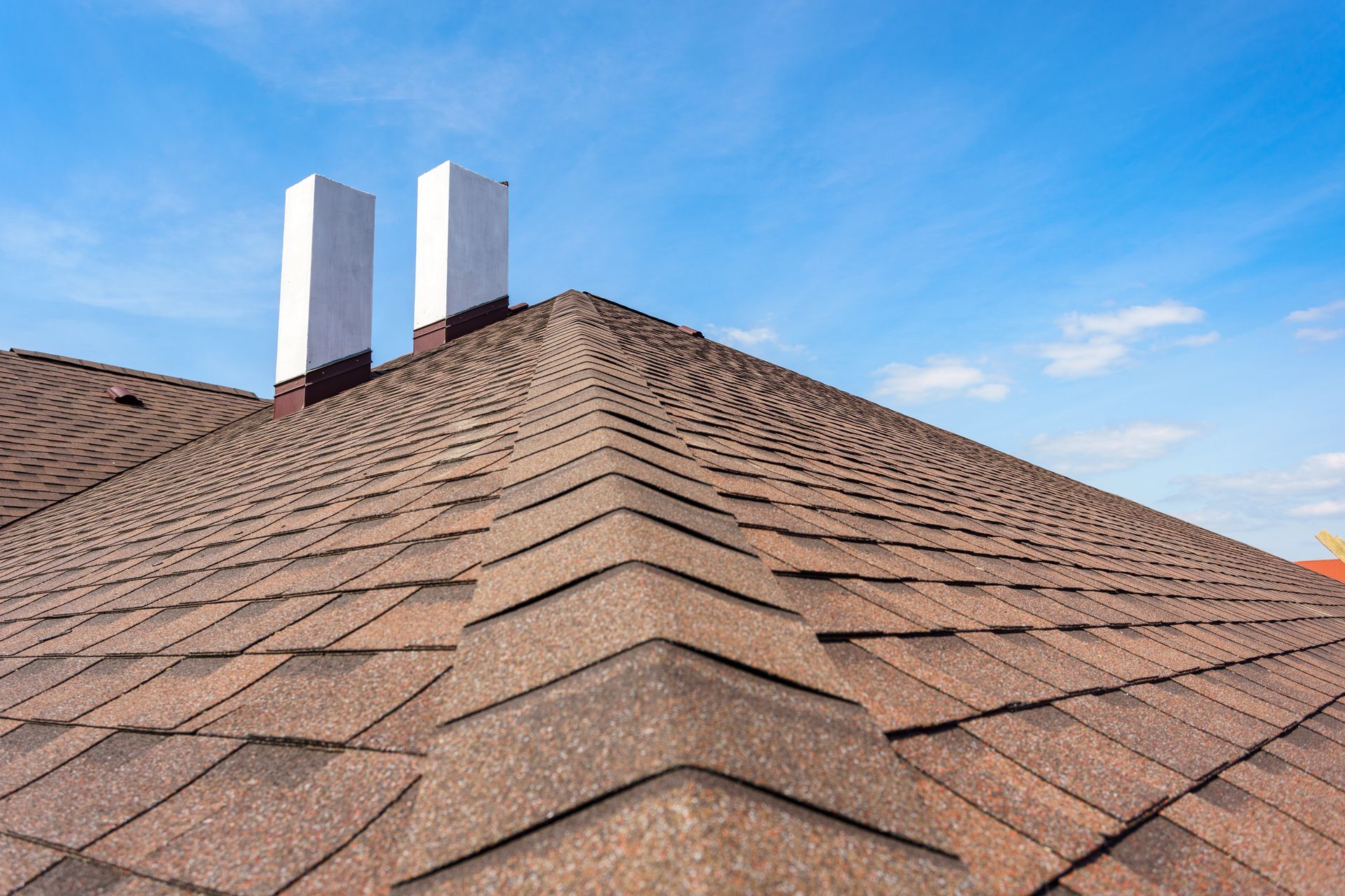 Rooftop with brown shingle tiles and two white chimney vents under a blue sky