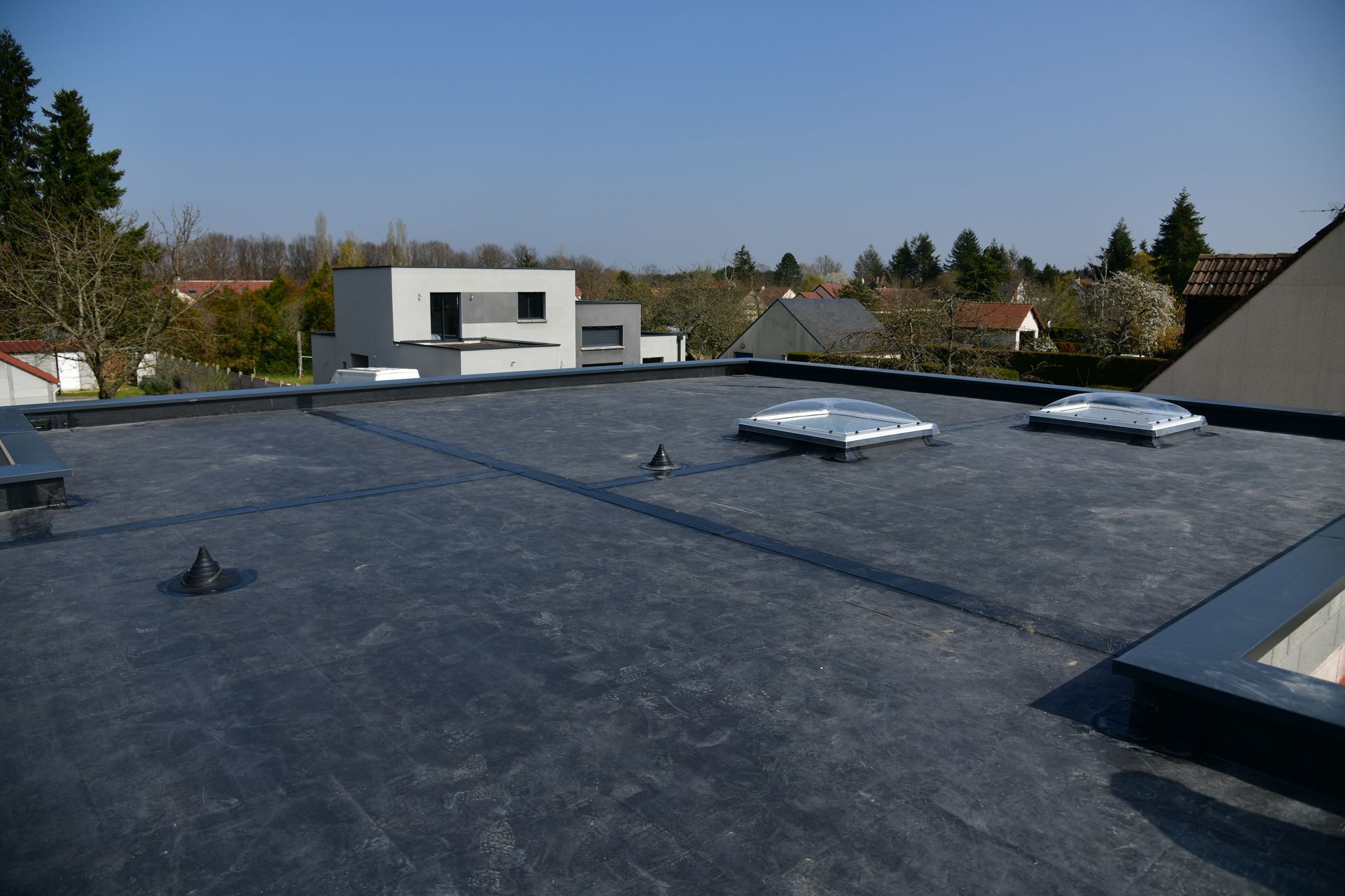 Flat dark rooftop with skylights and vents, overlooking houses under a clear blue sky.