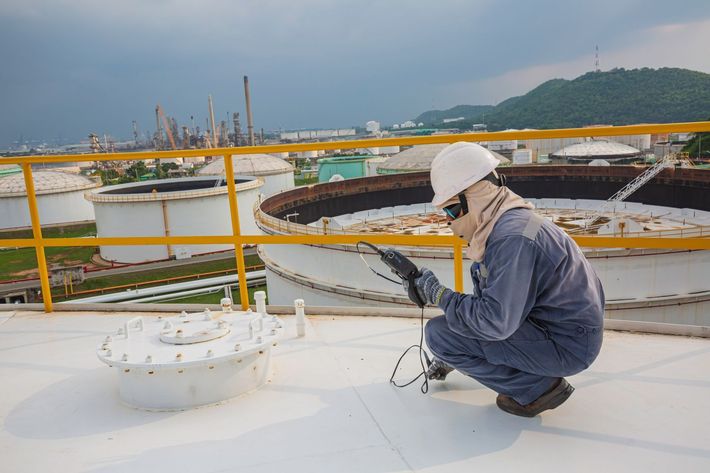 Worker in white hard hat crouches on a rooftop near industrial tanks and railings under cloudy skies