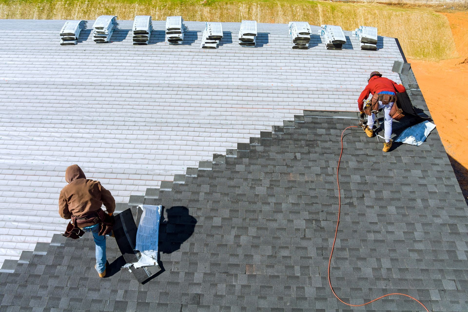 Two roofers working on a dark shingle roof beside a white roof with parked cars in the distance