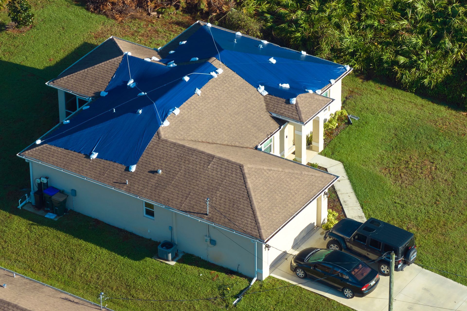 Aerial view of a house with a blue tarp-covered roof and parked vehicles in the driveway