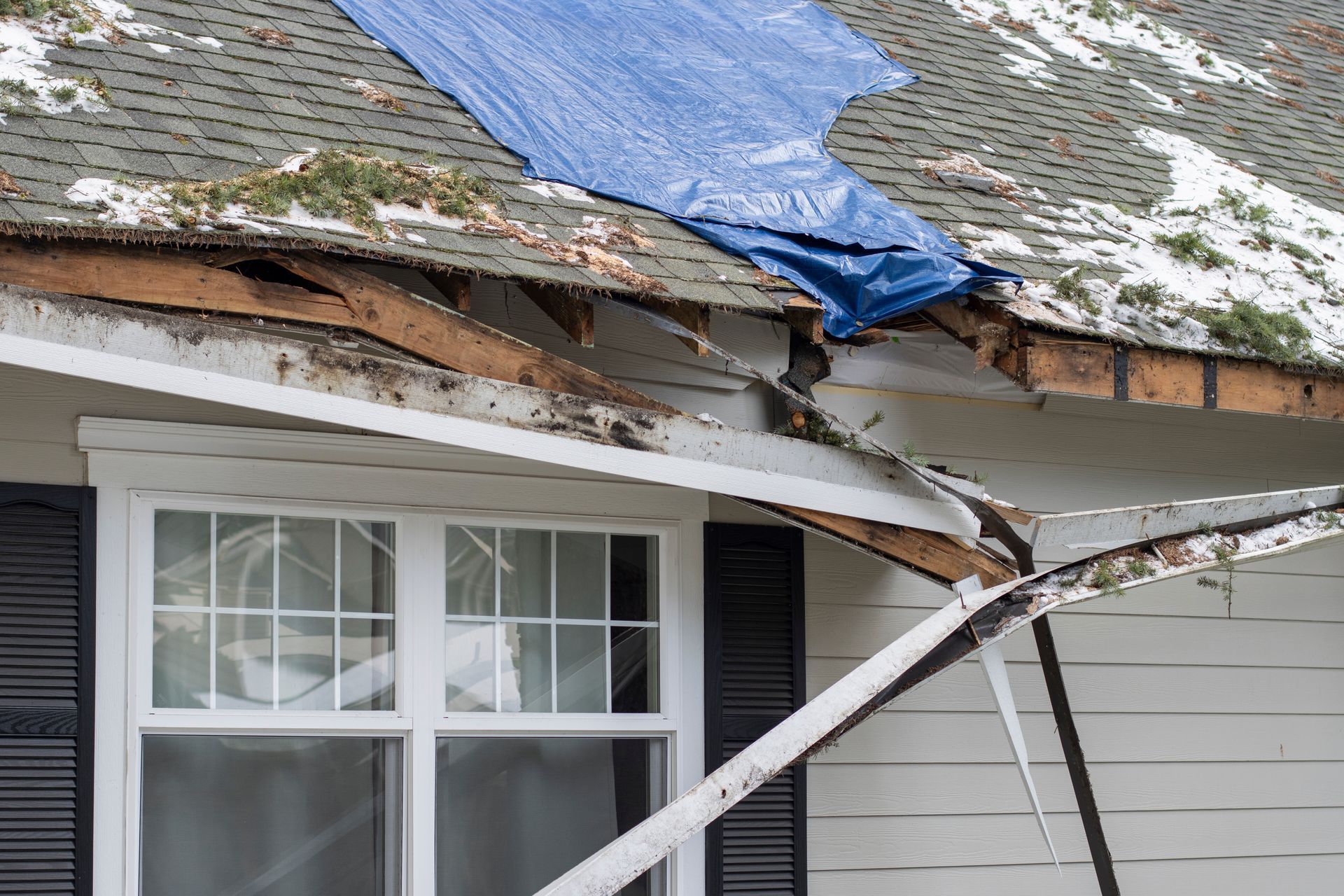 Damaged house roof with a blue tarp and broken white trim above a window