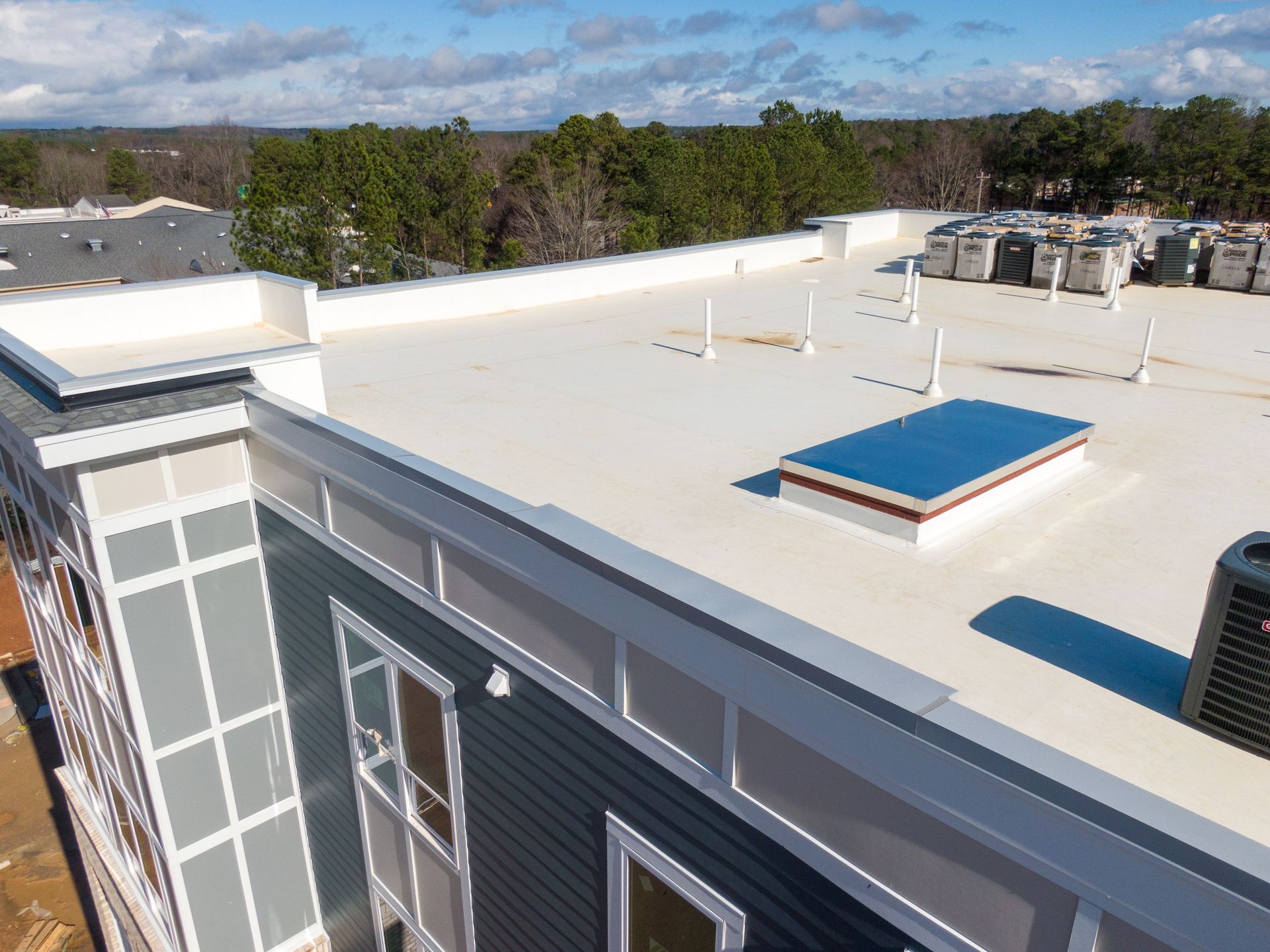 Rooftop with white flat surface, blue skylight, and vents on a building overlooking trees and sky.