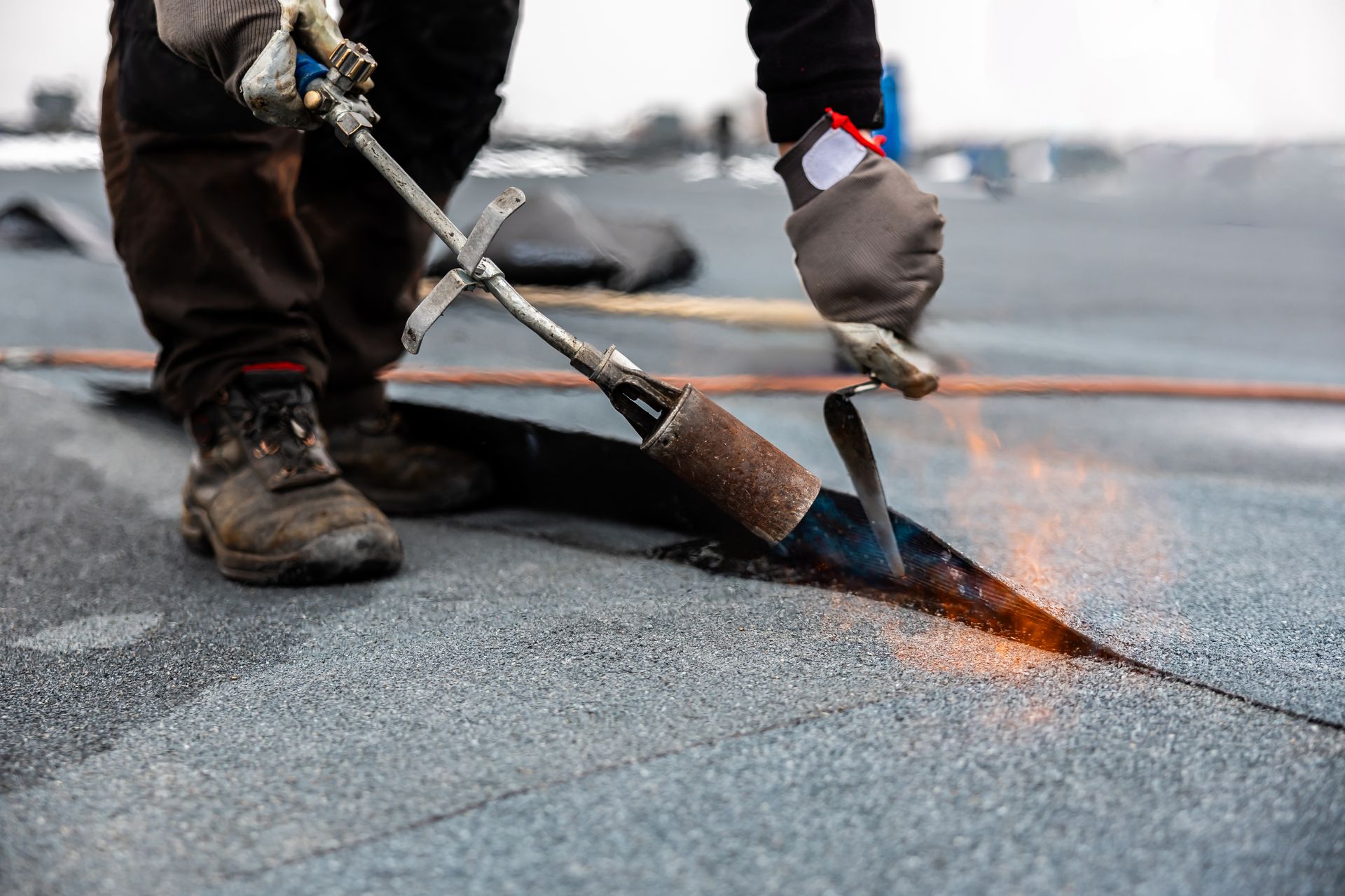Worker using a torch to seal roofing seam on a flat roof, with sparks and orange flame.